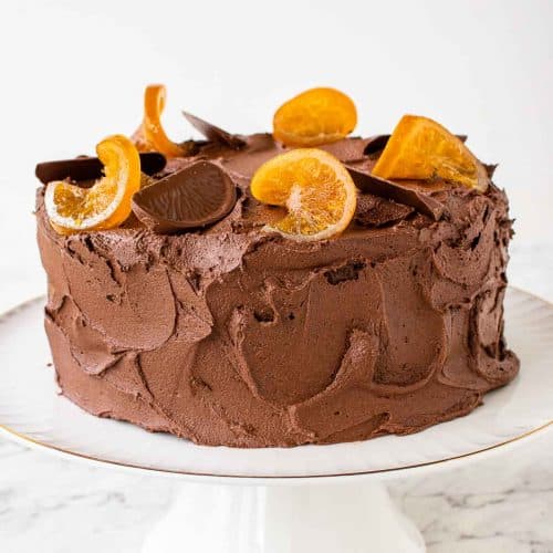 brown frosted cake on white cake stand with orange slices decoratively placed on top.