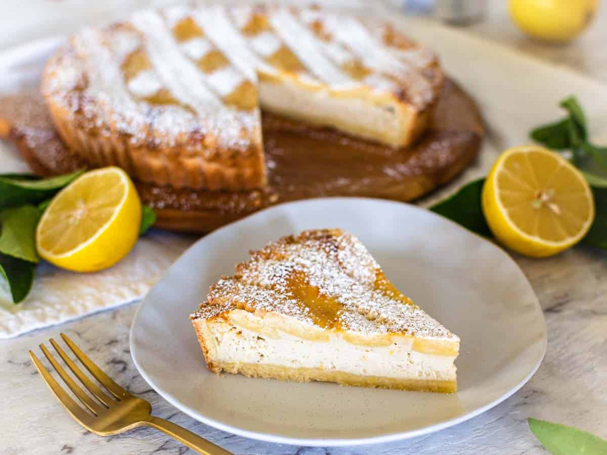 Slice of pie on white plate with whole pie in the background and halves lemons scattered around.