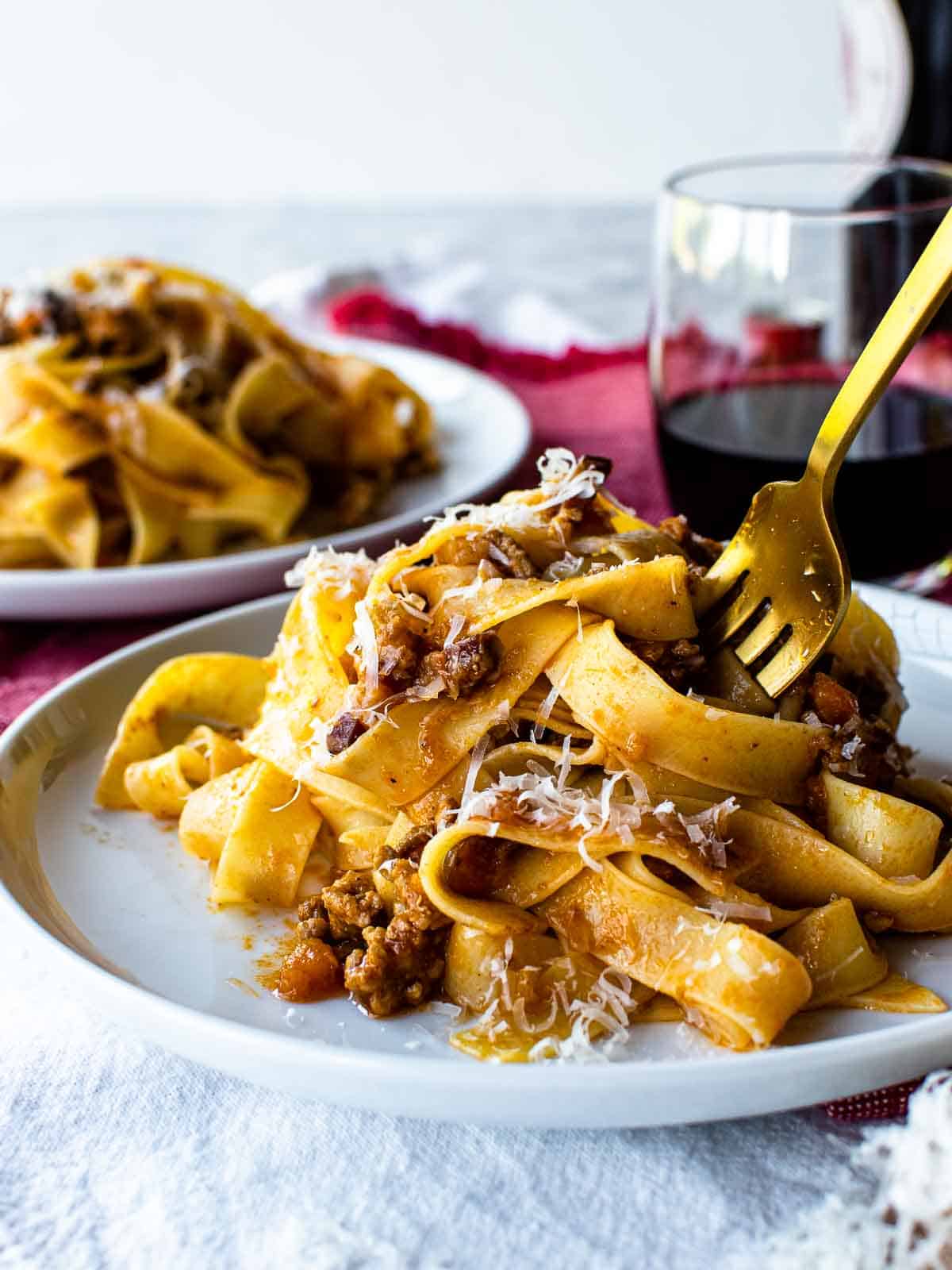 White plate with tagliatelle alla bolognese pasta on it and a gold fork in the pasta.