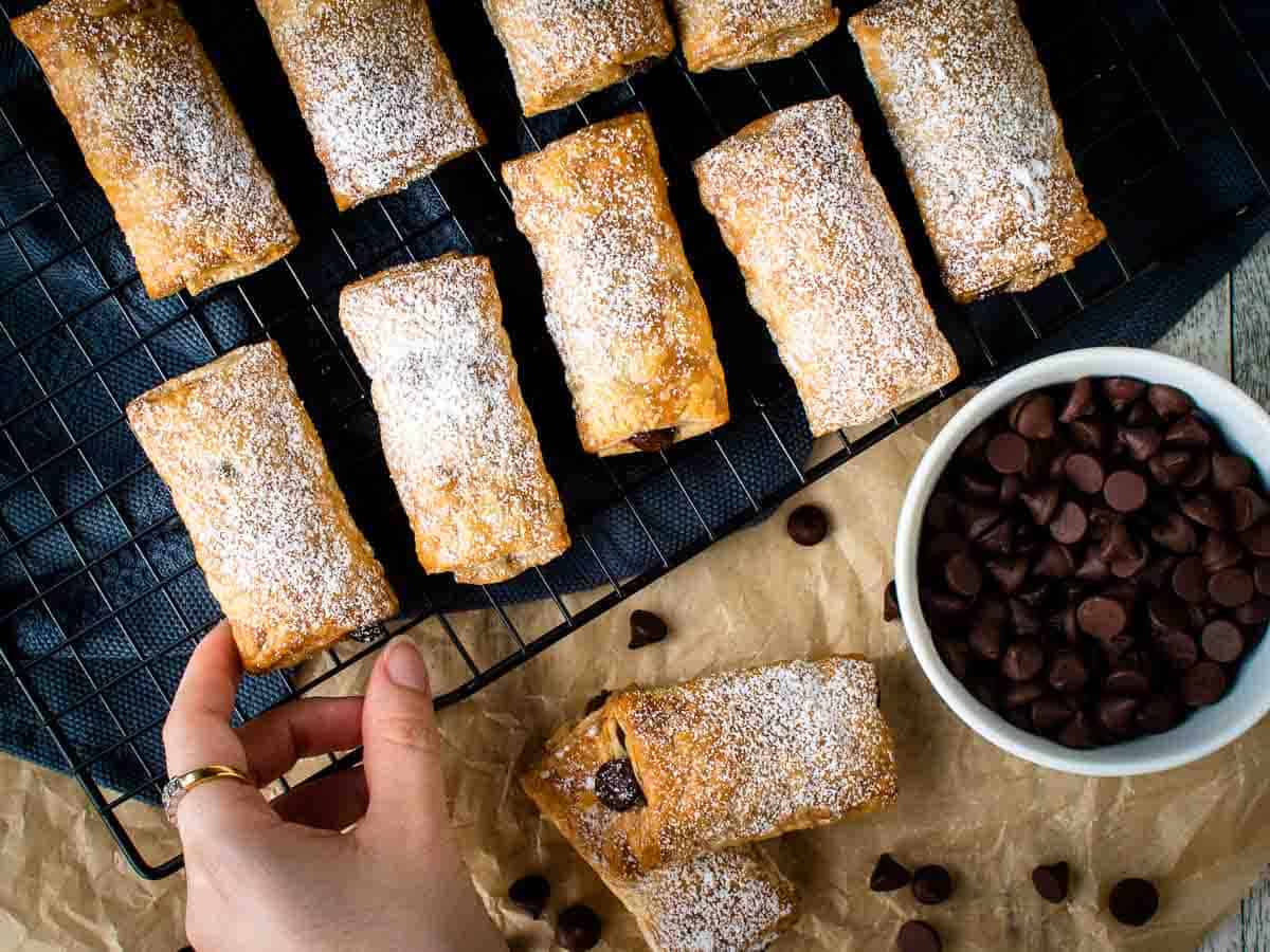 Rectangle pastries on a black wire rack with a hand choosing one, chocolate chips scattered around.