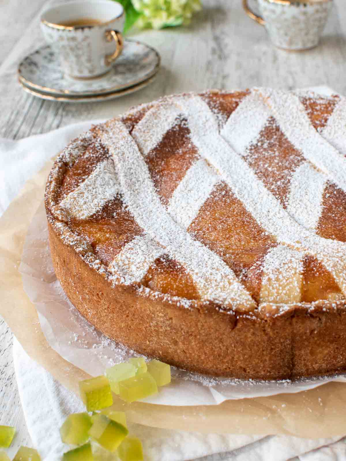 A round high Pastiera pie with lattice top dusted with powdered sugar, espresso in the background.