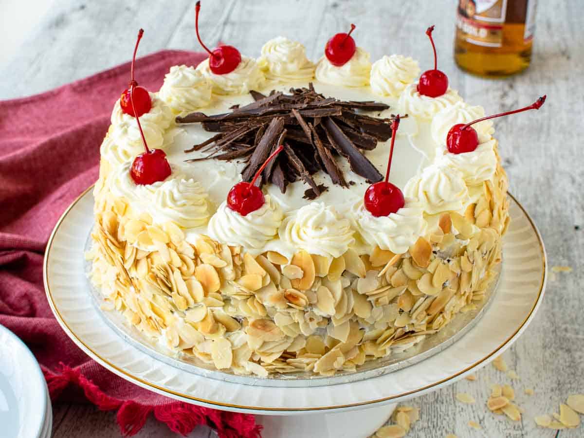 Cake topped with cream, cherries and chocolate and sides covered in sliced almonds.