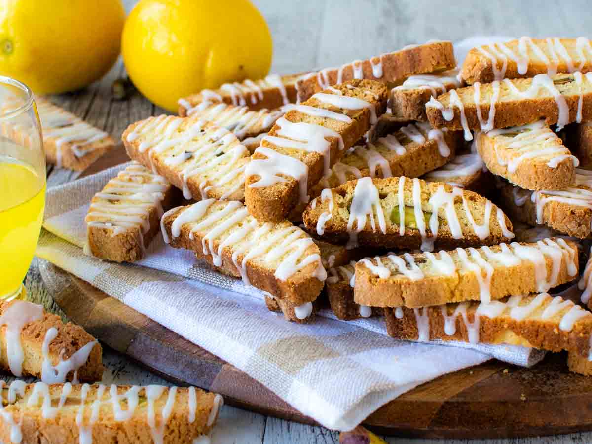 Pile of biscotti drizzled with white icing on a checked cloth with lemons in the background.