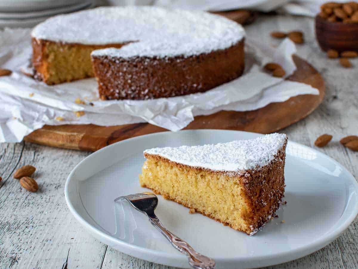 Slice of almond cake on a white plate with whole cake in the background.