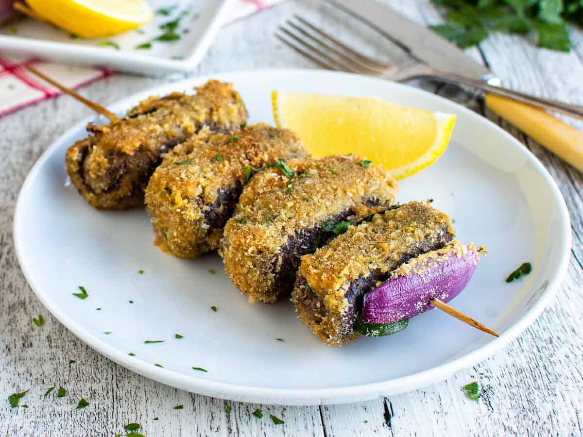 Four breaded beef rolls on wooden skewer on a white plate.