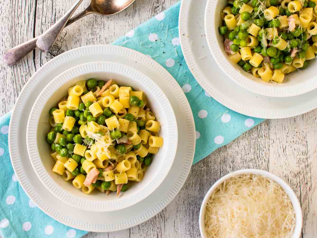 A white bowl filled with short pasta noodles and green peas viewed from above.