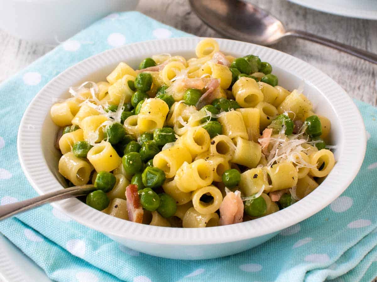 Close up of a white bowl filled with cooked small pasta, cubes of bacon and green peas.