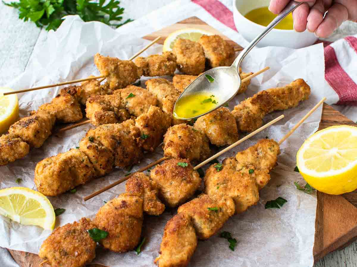 Chicken Skewers on white baking paper with a spoonful of melted butter dripping onto them.