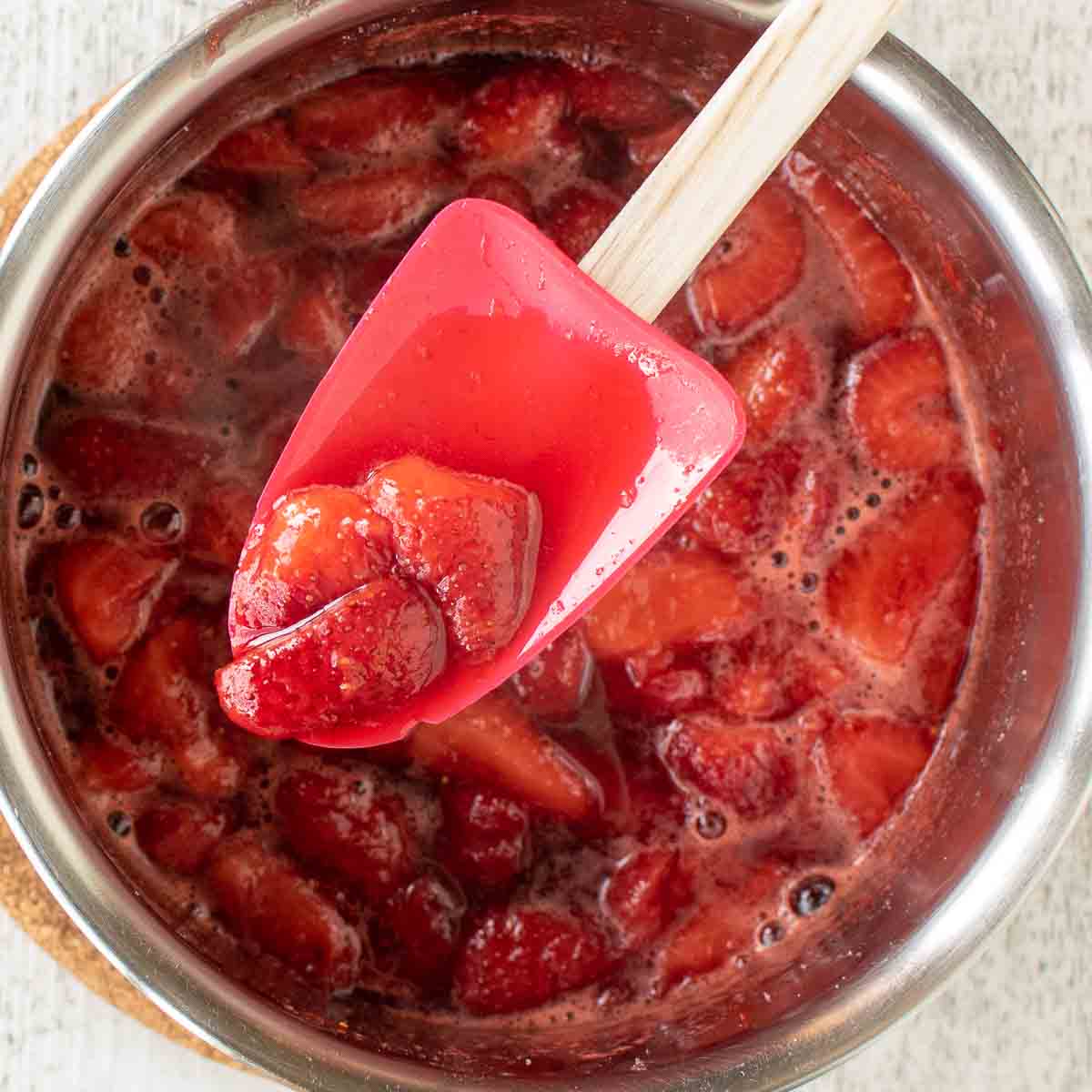 A spoonful of cooked strawberries on a red spatula viewed from above and held over a pot of cooked strawberries.
