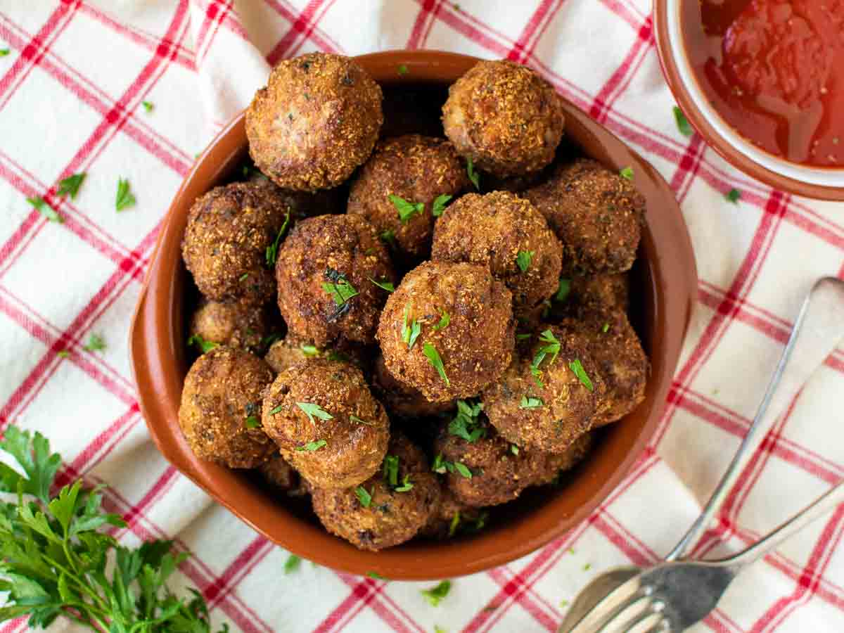 Brown meat balls piled onto a plate on a red and white checked tablecloth.