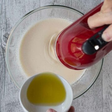 Adding olive oil to beaten eggs and sugar using hand held red electric mixer.