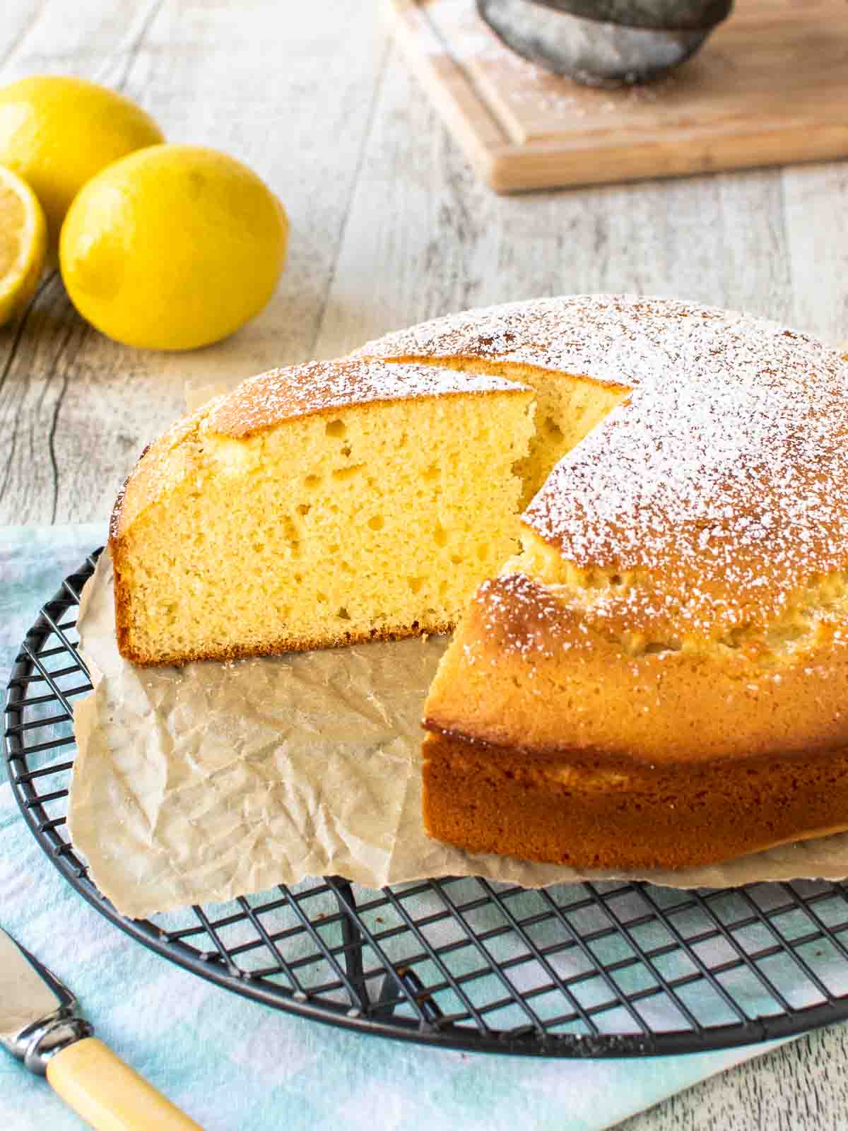 Italian Lemon Cake with a slice removed on a black wire cooling rack with lemons in the background.