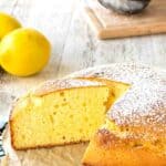 Italian Lemon Cake with a slice removed on a black wire cooling rack with lemons in the background.