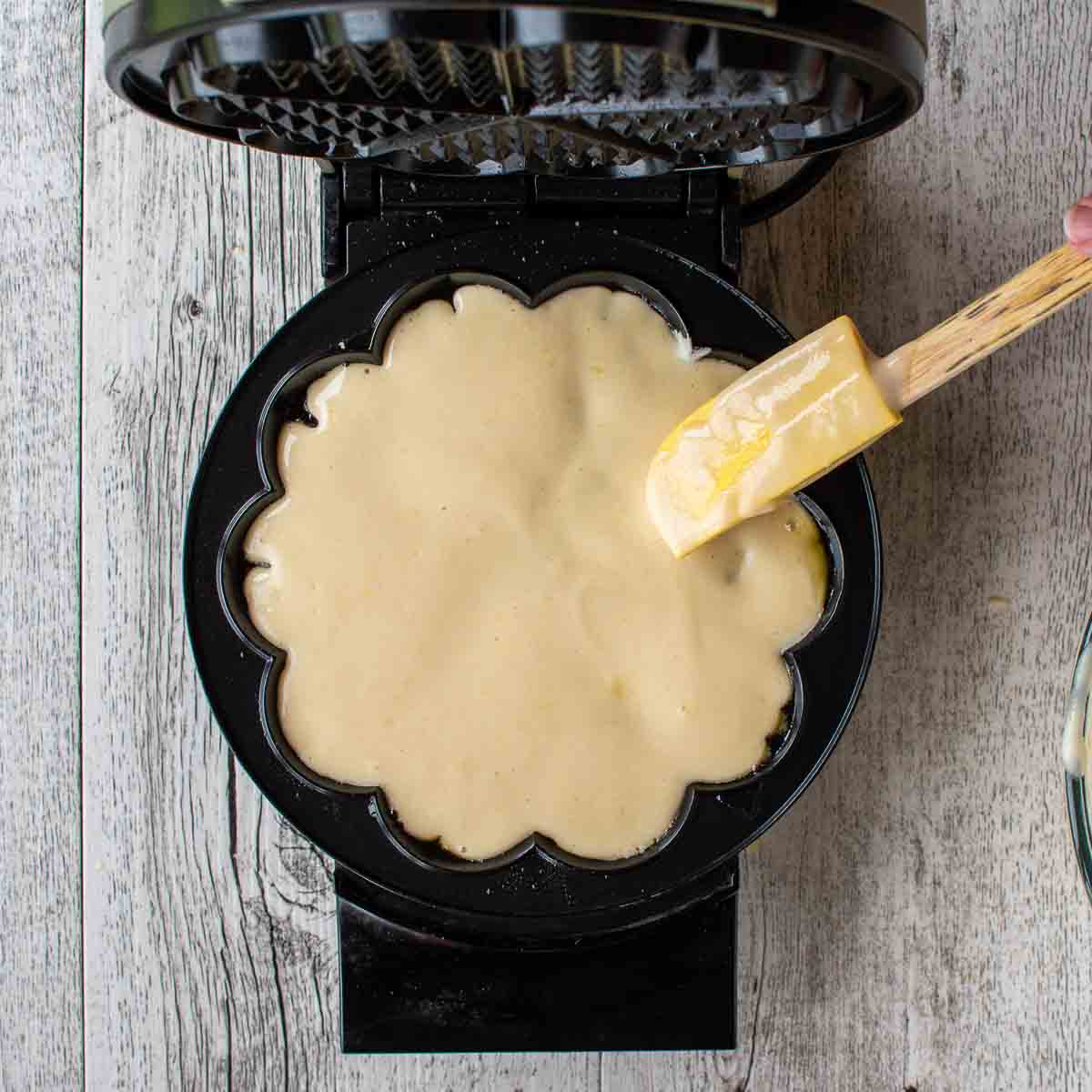 Waffle batter being spread on waffle iron with a yellow spatula viewed from above.