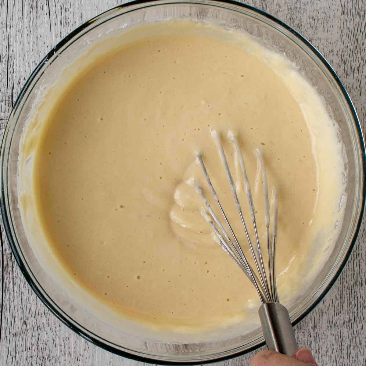 Thick waffle batter and a whisk in a glass bowl viewed from above.