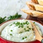 Garlic and herb cheese spread in a red bowl, garnished with chopped chives.