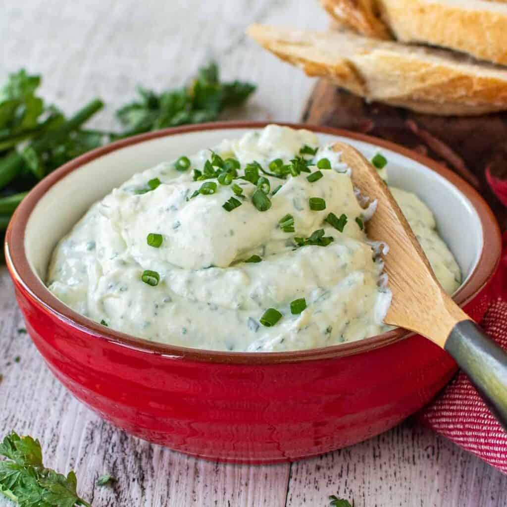 Garlic and herb cheese spread in a red bowl, garnished with chopped chives.