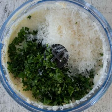 Chopped herbs and grated Parmesan cheese in a mini food processor viewed from above.