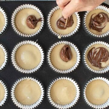 Yellow batter in cupcake papers. Some topped with Nutella spread and being swirled around with a toothpick.
