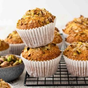 Two pistachio muffins stacked on a black wire rack with more muffins in the background.