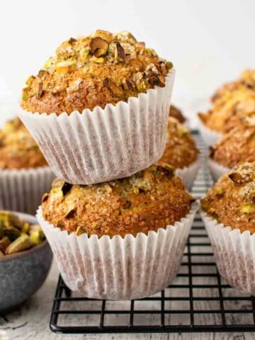 Two pistachio muffins stacked on a black wire rack with more muffins in the background.
