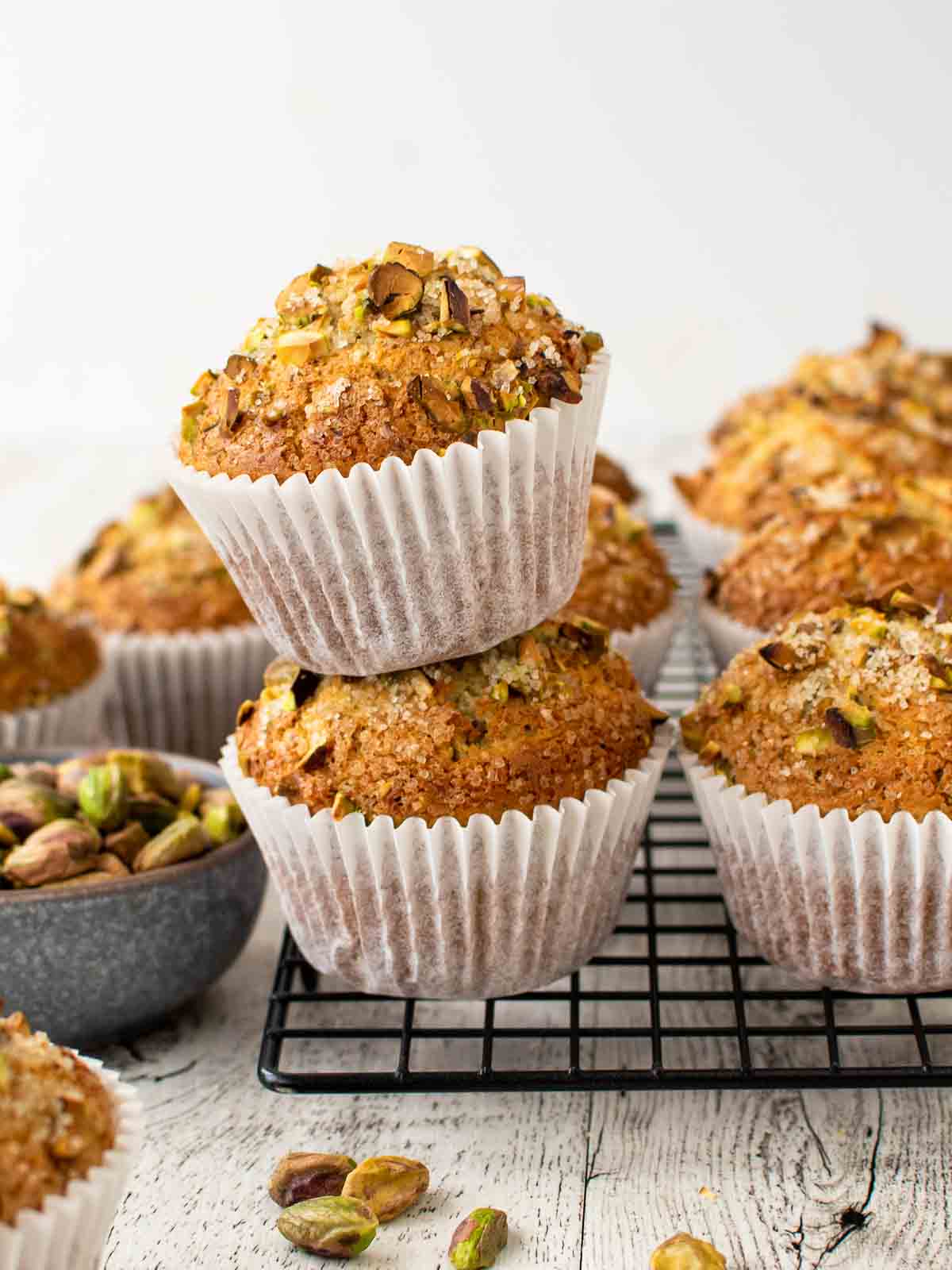 Two pistachio muffins stacked on a black wire rack with more muffins in the background.