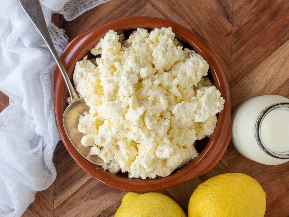 Overhead view of ricotta cheese in a terracotta bowl surrounded by milk bottle, lemons and cheesecloth.