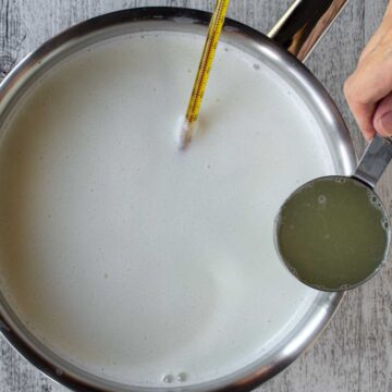 Milk in a saucepan with measuring cup of lemon juice about to be poured in.