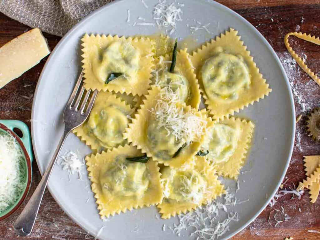 A white plate on a wooden table holds several pieces of ravioli topped with grated cheese. A fork is placed on the left side of the plate. A chunk of cheese, a bowl with more grated cheese, and scattered cheese shavings surround the plate.