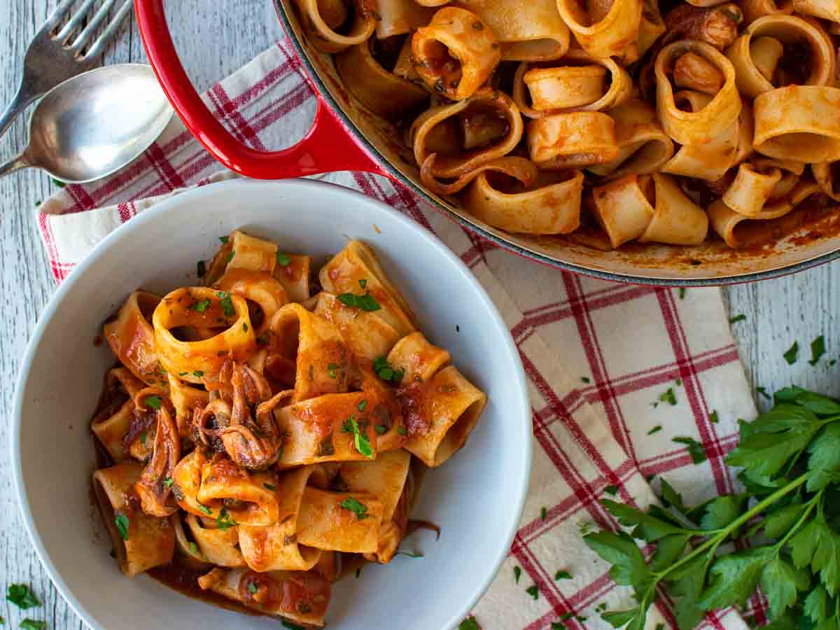 Overhead view of a tomato and calamari pasta in a bowl with a pan full nearby.