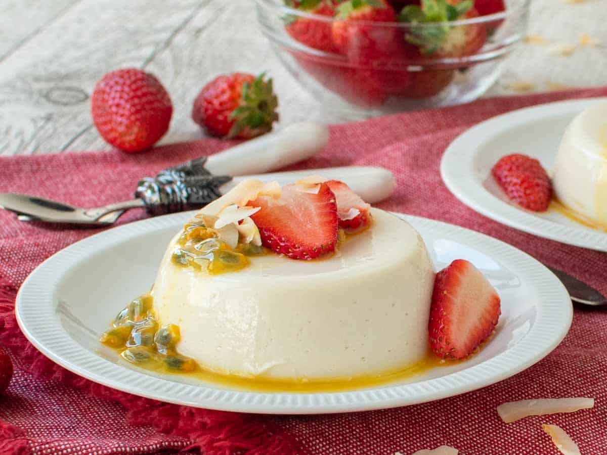 Close up of white unmolded pudding on a white plate topped with sliced strawberries and passion fruit.