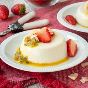 Coconut panna cotta on a white plate topped with sliced strawberries and passion fruit with more in the background.