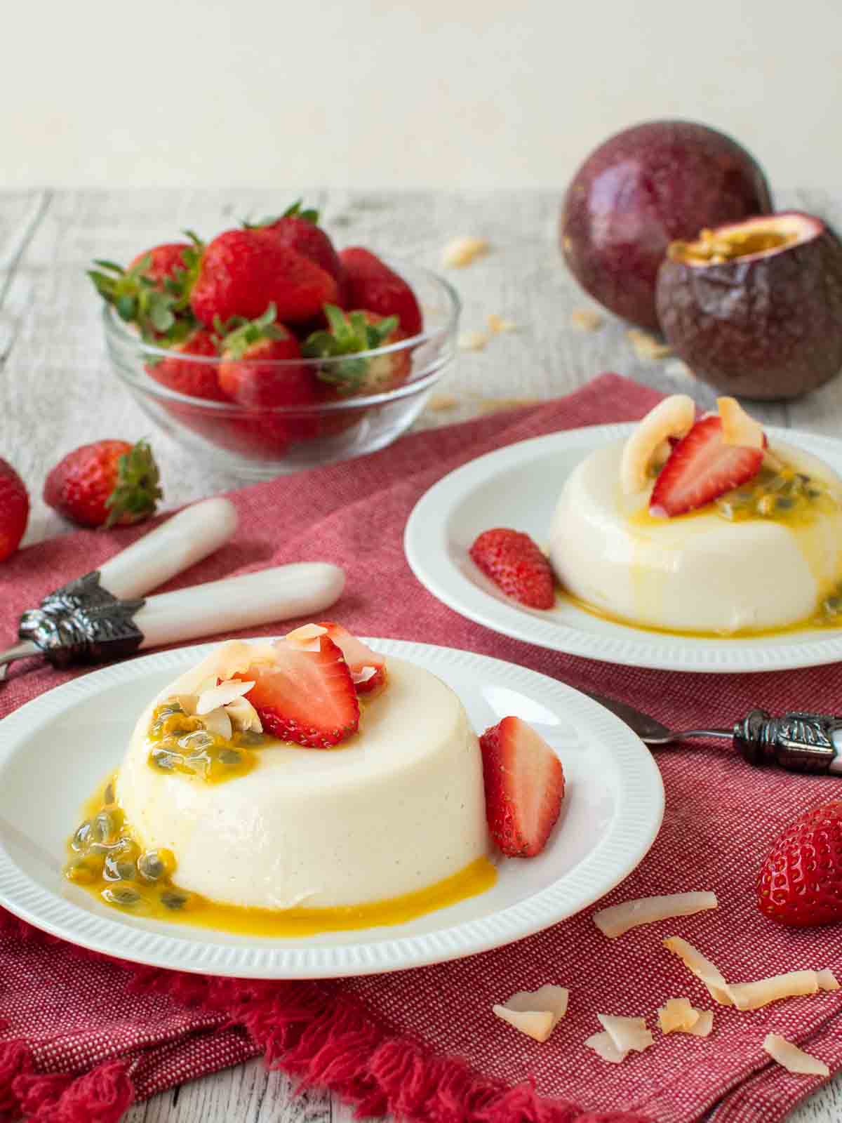 Coconut panna cotta on a white plate topped with sliced strawberries and passion fruit with more in the background.