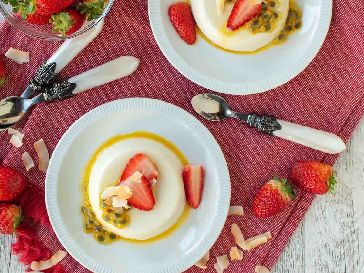 Overhead view of unmolded panna cotta with coconut milk topped with strawberries and passionfruit on a white plate.