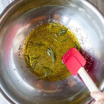 Oil and herbs in a stainless steel bowl with a red spatula viewed from above.