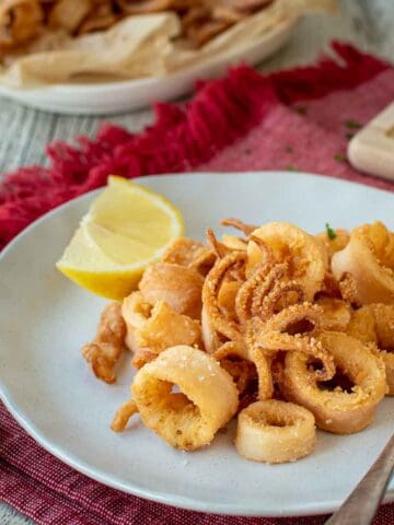 Calamari Fritti on a white plate with a wedge of lemon on the side and more calamari and lemon in the background.