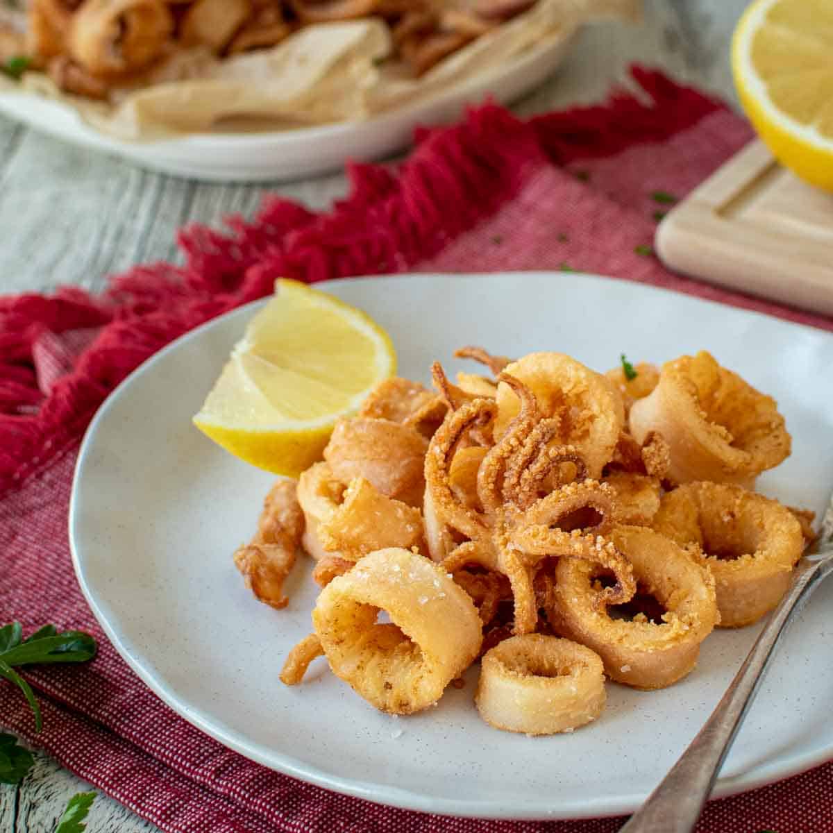 Calamari Fritti on a white plate with a wedge of lemon on the side and more calamari and lemon in the background.