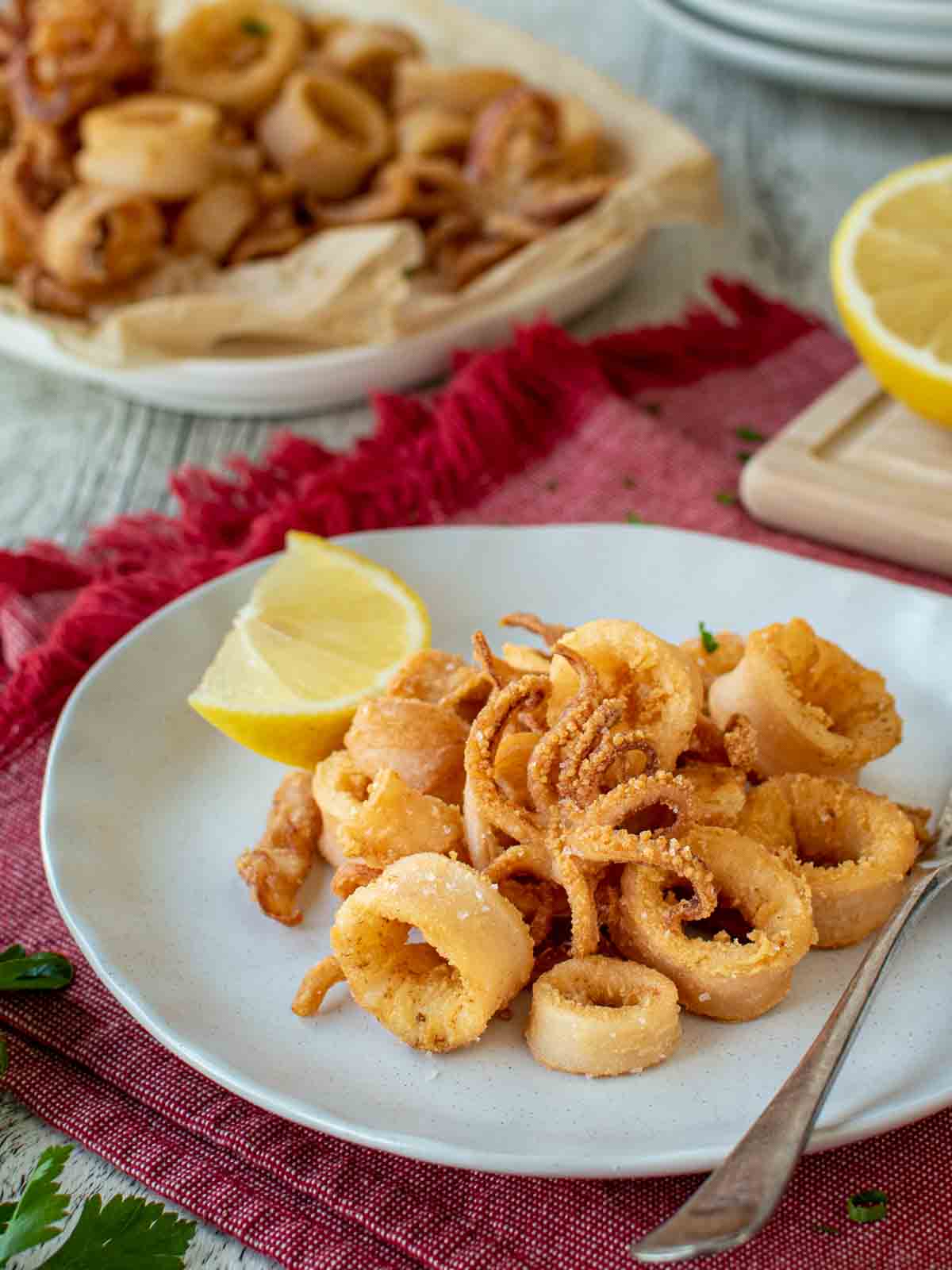 Calamari Fritti on a white plate with a wedge of lemon on the side and more calamari and lemon in the background.