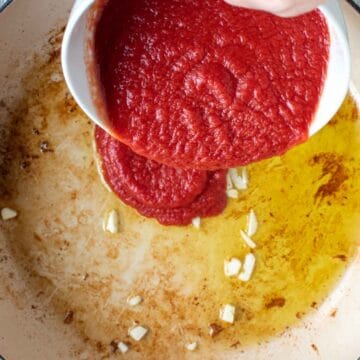 Tomato puree being poured into a pan with containing oil and some chopped garlic.