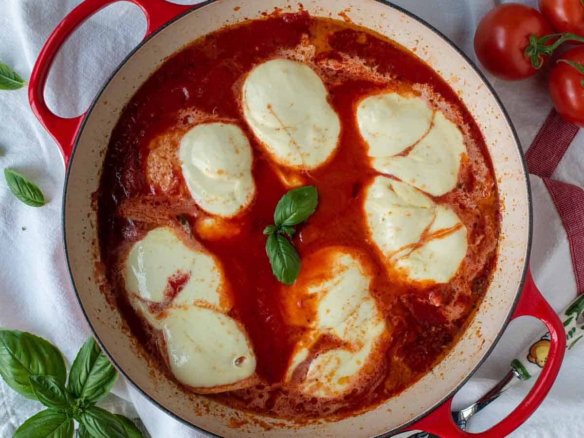 Overhead view of cheese topped chicken breasts in a tomato puree in a red pan.