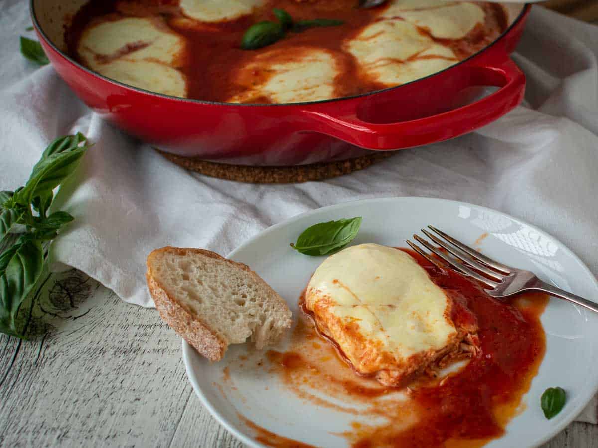 Cheese topped chicken in tomato puree on a white plate.