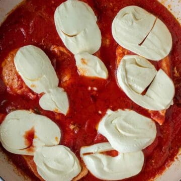 Slices of cheese on chicken breasts in a pan of tomato puree.