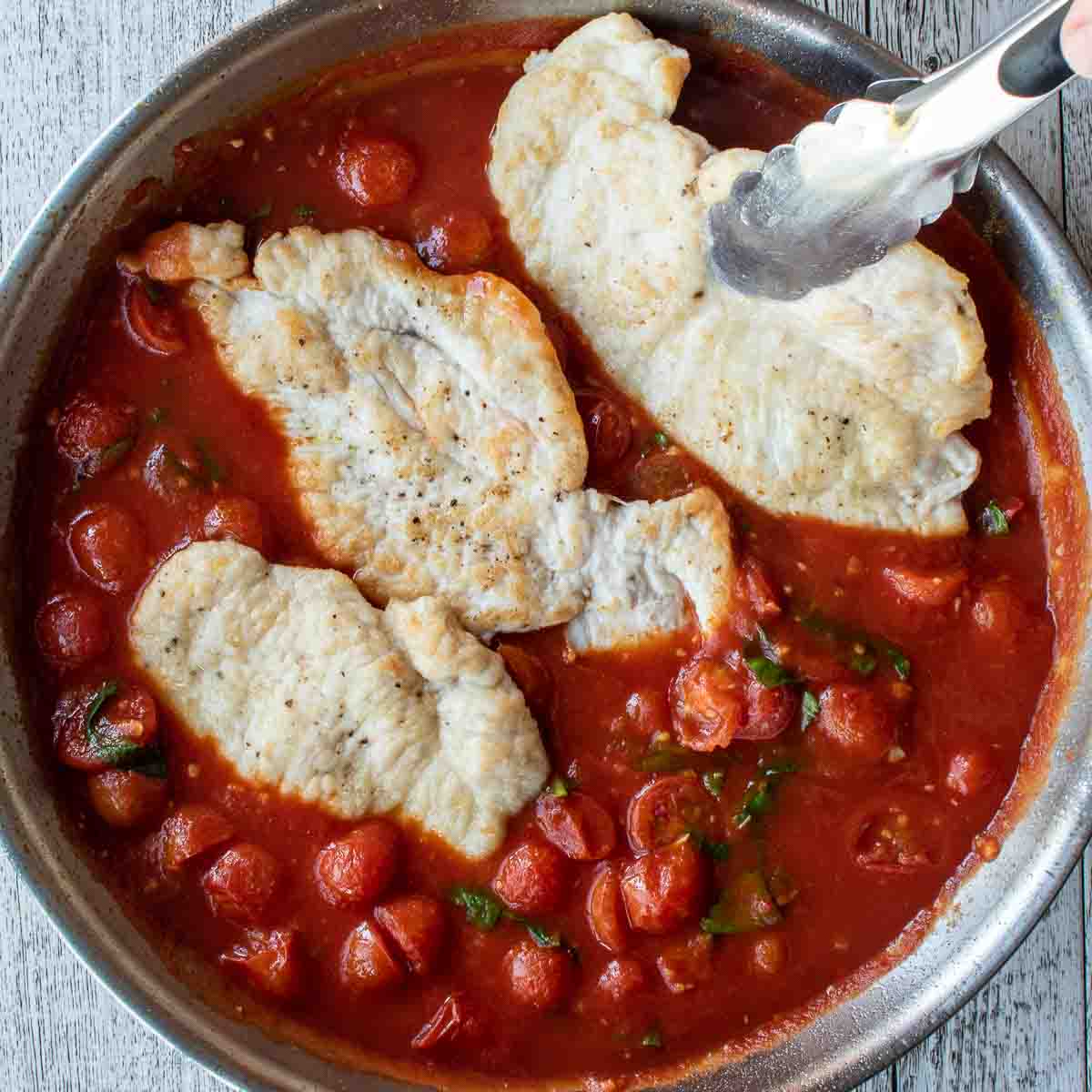 Chicken cutlets being added to a tomato sauce in a stainless steel pan.