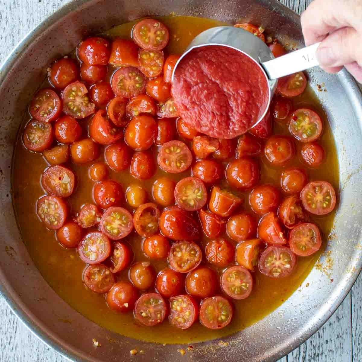 Tomato puree being poured into a pan of halved cherry tomatoes and more reddish liquid.