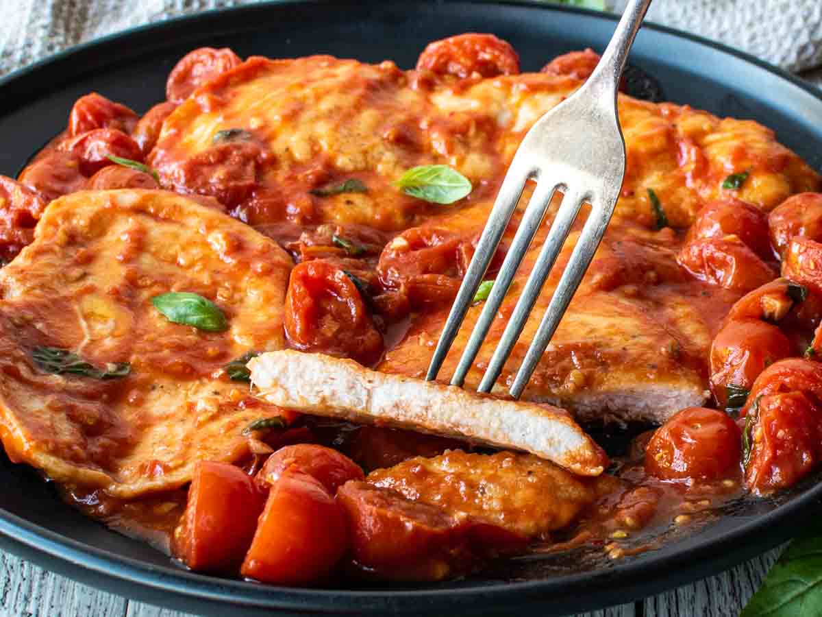 A fork with a piece of chicken breast on it and more chicken and tomato sauce in the background.