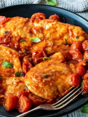 Chicken pomodoro with cherry tomatoes and fresh basil scattered around on a black plate.