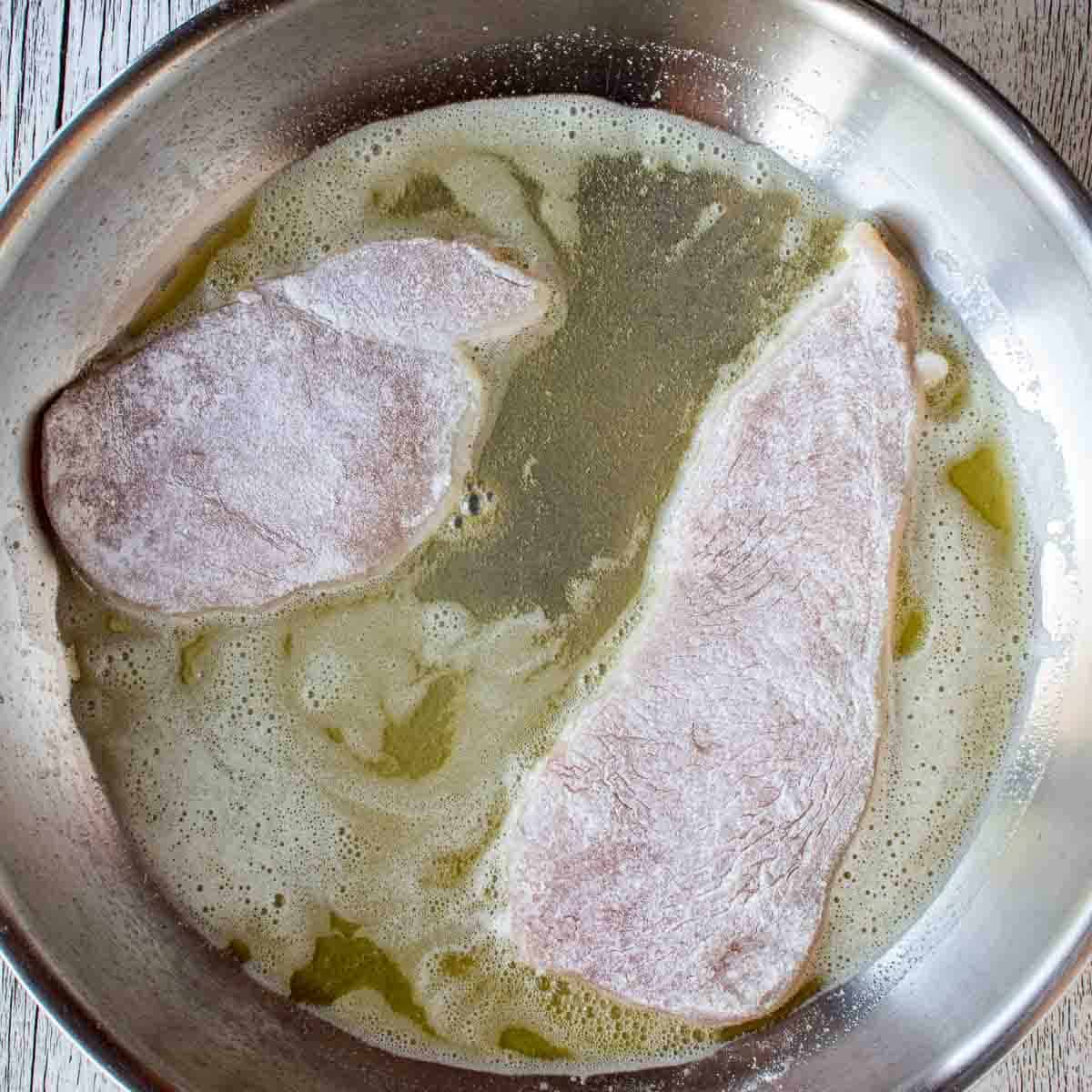 Flour coated chicken cutlets frying in oil in a stainless steel pan.