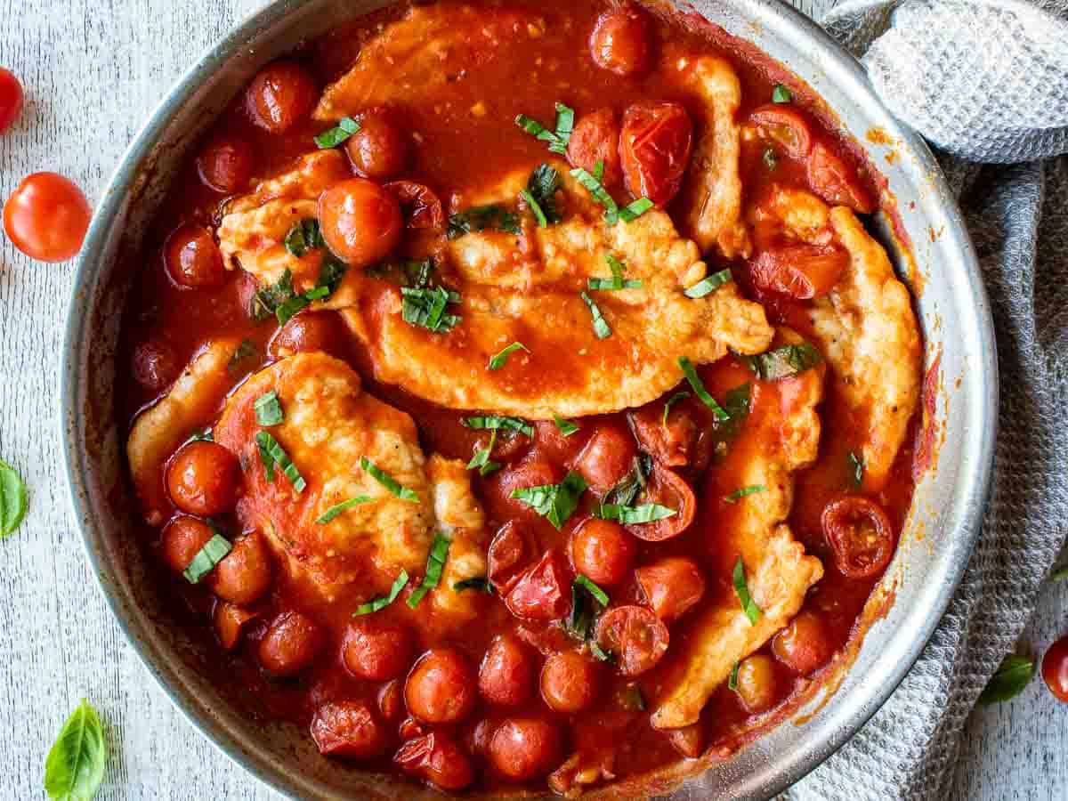 Chicken cutlets in a cherry tomato sauce in a stainless steel pan.