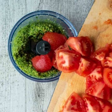 Chopped tomatoes being scraped into a mini food processor filled with a smooth green paste.