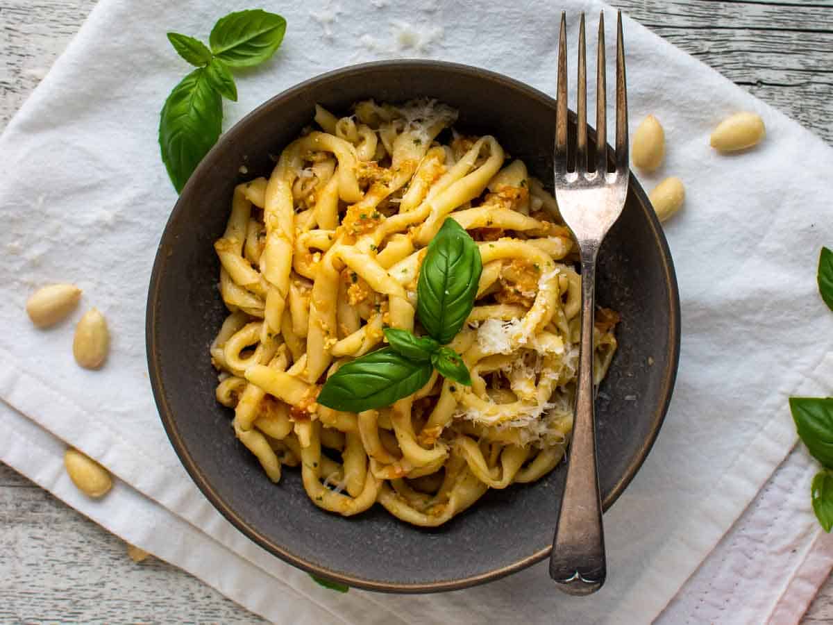 A grey bowl filled with handmade, twirled pasta with a reddish sauce, topped with a sprig of basil and a fork balance on the edge of the bowl viewed from above.